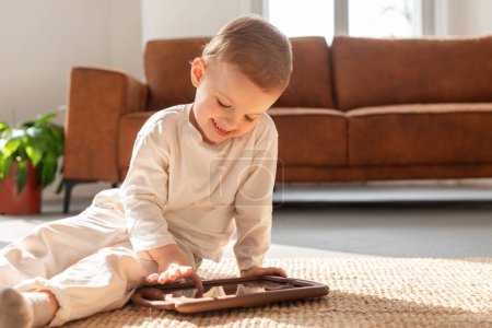 Téléchargez les photos : Un jeune garçon est assis sur le sol, absorbé par le jeu sur une tablette. Il se concentre sur l'écran, tapant et glissant avec concentration - en image libre de droit