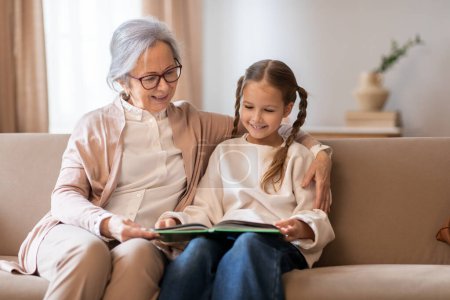An older woman and a young girl are seated together on a couch, engrossed in a book that they are reading together. Grandmother hand rests on the book, guiding the young girl