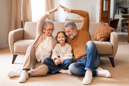 A joyful elderly couple sits on the floor with their young granddaughter between them, smiling and creating roof shape with their hands over the girls head