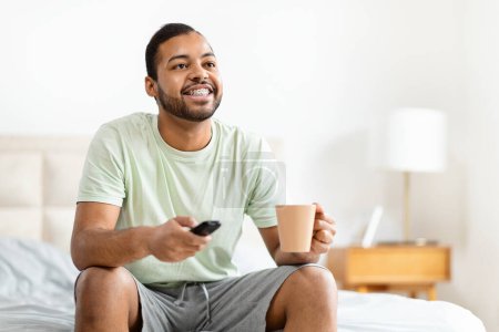 A cheerful African American man in casual clothing sits comfortably on his bed holding a coffee mug in one hand and a remote control in the other