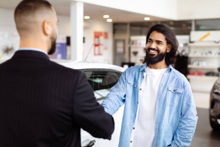 Two men are standing in a car showroom, shaking hands in agreement. The men appear serious and focused on finalizing a deal for a car purchase