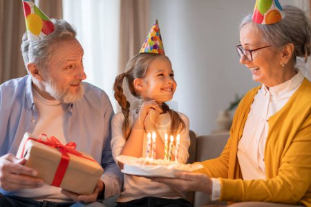 Una joven vestida con un sombrero de cumpleaños sonríe brillantemente mientras sus abuelos, también vistiendo sombreros de fiesta festivos, le regalan un pastel de cumpleaños adornado con velas encendidas