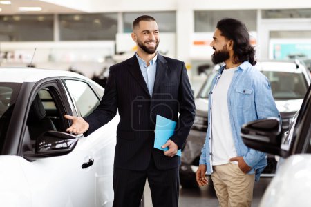 A smartly dressed car salesman, with a friendly demeanor, stands next to a shiny new car in a well-lit vehicle showroom, engaging with a potential customer Indian guy who is holding a brochure.