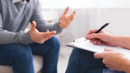 A person is seated in a neutral-colored office space, engaging in a discussion with another individual who is taking notes on a pad, cropped