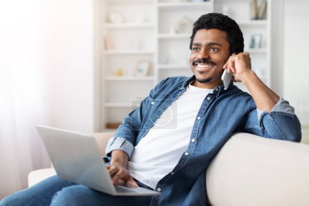 A cheerful African American man sits comfortably on a white sofa with a laptop on his lap, engaging in a conversation on his smartphone