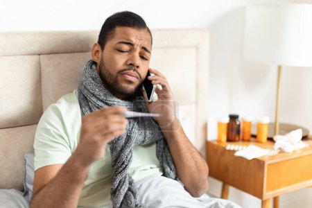 Foto de Un joven afroamericano está sentado en la cama con una bufanda gris envuelta alrededor de su cuello, apareciendo mal mientras hace una llamada telefónica, hablando con el médico a distancia. - Imagen libre de derechos
