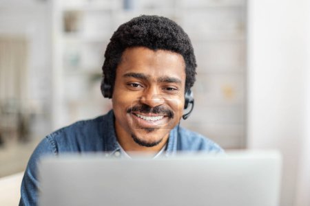 A smiling African American man wearing a headset sits in front of a laptop, likely providing customer assistance. The office environment is bright and airy, closeup
