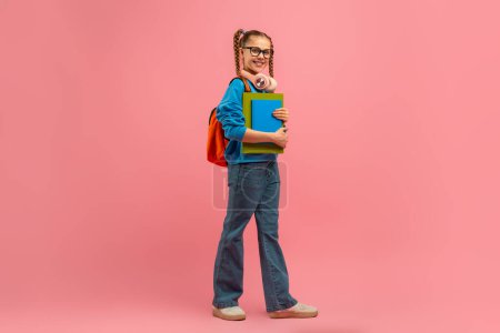 Foto de Una joven de pie con una mochila colgando de su hombro, sosteniendo un libro en sus manos. Parece centrada en el libro, posiblemente a punto de embarcarse en un viaje o regresar de la escuela.. - Imagen libre de derechos