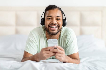African American man is pictured wearing headphones and engaging with a cell phone. He seems focused on the screen, possibly listening to music or having a phone conversation.