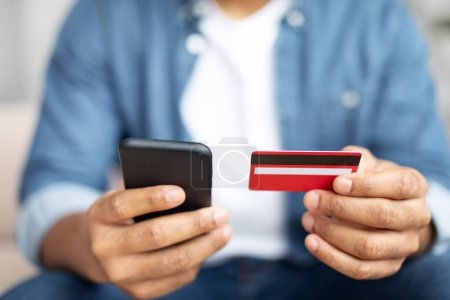Téléchargez les photos : Coupé d'homme afro-américain tenant une carte de crédit dans une main et un téléphone portable dans l'autre, effectuant un achat ou une transaction en ligne. Opérations financières modernes utilisant la technologie. - en image libre de droit