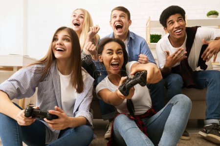 Eine Gruppe multiethnischer Freunde hat sich um eine Couch versammelt und spielt gemeinsam Videospiele. Sie sitzen eng beieinander, halten Spielcontroller in der Hand und konzentrieren sich auf den Bildschirm..