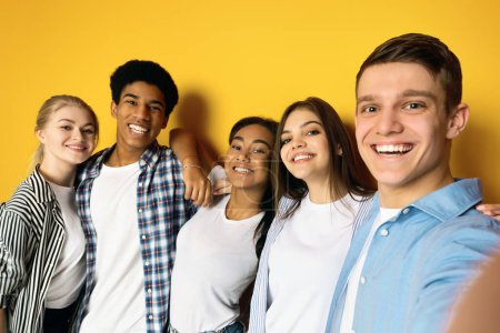 Eine fröhliche Gruppe von fünf multiethnischen Teenagern posiert eng beieinander in einer freundlichen Umarmung, macht Selfie auf gelbem Studiohintergrund