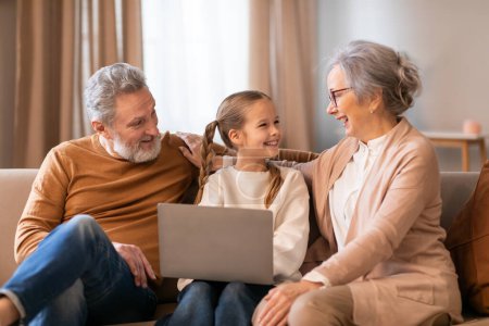 Una joven alegre está sentada en un acogedor sofá entre sus abuelos, con un portátil abierto frente a ellos. Están ocupados en un momento conmovedor.