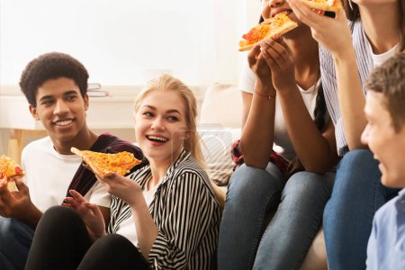 Photo for A cheerful group of young friends sits comfortably on the floor and sofa in a well-lit living room, sharing slices of pizza and engaging in lively conversation - Royalty Free Image