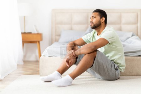 Ein junger afroamerikanischer Mann sitzt auf dem Boden neben dem Bett in einem sonnendurchfluteten Schlafzimmer, er wirkt aufgebracht, frustriert, deprimiert, Kopierraum