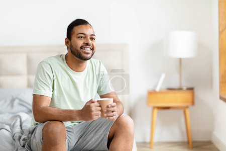Téléchargez les photos : Afro-Américain est assis sur un lit, tenant une tasse de café dans ses mains. Il semble détendu et contemplatif comme il aime sa boisson du matin, copier l'espace - en image libre de droit