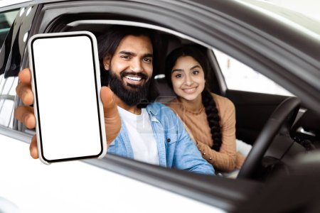 Téléchargez les photos : Un Indien barbu étend son bras par la fenêtre d'une voiture, tenant un smartphone avec un écran vide vers la caméra, éventuellement pour un affichage interactif ou à des fins publicitaires. - en image libre de droit