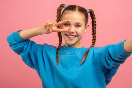 A cheerful young girl in a bright blue sweater is striking a playful pose with one hand, flashing the peace sign near her eye, while her other arm extends outward as if reaching for the camera.