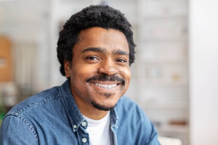 Foto de Un joven y alegre afroamericano con bigote y pelo rizado se viste casualmente con una camisa de mezclilla. Da una sonrisa conmovedora, interior del hogar, primer plano - Imagen libre de derechos