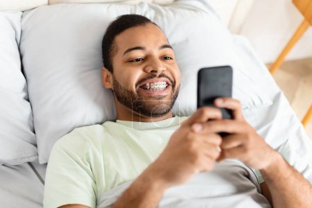Foto de El hombre afroamericano está acostado en una cama, sosteniendo un teléfono celular en su mano. Parece estar desplazándose por la pantalla, posiblemente revisando mensajes o navegando por las redes sociales.. - Imagen libre de derechos