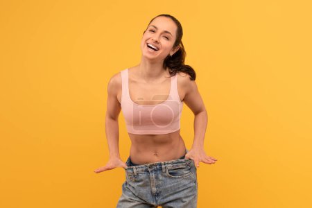 Eine strahlende junge Frau zieht selbstbewusst an ihrer überdimensionalen Jeans, um ihre erfolgreiche Gewichtsabnahme, ihr Glück und ihren Stolz zu präsentieren, während sie vor einem leuchtend gelben Hintergrund posiert..