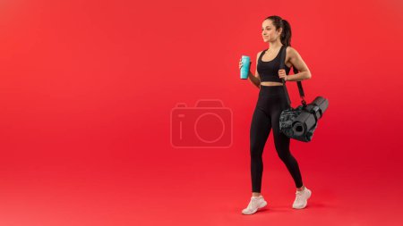 Photo for A fit woman dressed in black sportswear confidently strides forward while holding a yoga mat and gym bag. Her white sneakers contrast with the bright red backdrop - Royalty Free Image