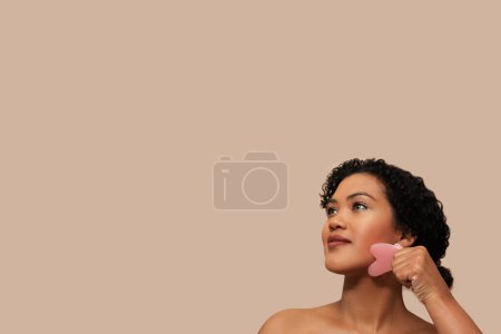 Foto de Mujer joven con el pelo rizado realiza un masaje facial utilizando una piedra de gua sha rosa, una herramienta de medicina tradicional china. Ella es capturada en medio del movimiento con la piedra sostenida contra su mandíbula, espacio de copia - Imagen libre de derechos
