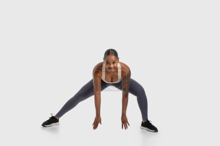 African American woman is seen performing a squat exercise on a white background. She is bending her knees while keeping her back straight, engaging her leg muscles. The white background
