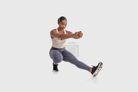 Mujer afroamericana se muestra haciendo un ejercicio en cuclillas sobre un fondo blanco llano. Ella está doblando sus rodillas y bajando sus caderas mientras mantiene su espalda recta y los brazos extendidos delante de ella