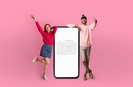Téléchargez les photos : Homme indien et femme européenne posent énergiquement avec des sourires, debout à côté d'un modèle surdimensionné d'un smartphone avec un écran vide - en image libre de droit