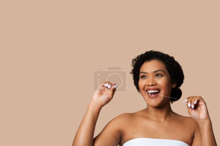 Photo for A cheerful woman with short, curly hair is captured mid-floss, taking care of her dental hygiene. She is wrapped in a towel, suggesting a setting possibly post-shower, copy space - Royalty Free Image