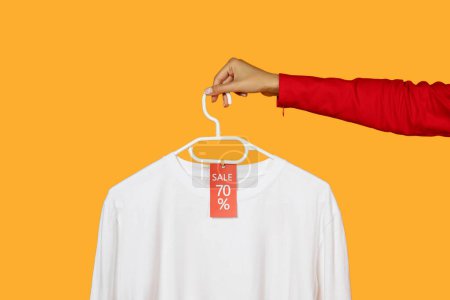Woman hand emerging from a red sleeve, holding a white hanger with a crisp white shirt on it. A red sale tag showing 70 percent is attached to the shirt, suggesting a retail discount offer