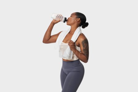 Mujer afroamericana en ropa deportiva sosteniendo una botella de plástico en sus labios, bebiendo agua aislada sobre fondo blanco