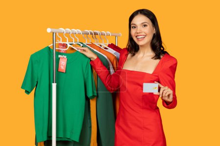 A woman is standing in front of a rack of clothes, holding a credit card in her hand. She appears to be shopping for clothing items at a store, making a payment with the credit card.
