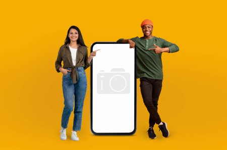 Téléchargez les photos : Un couple multiracial joyeux se tient à côté d'un écran de smartphone vide surdimensionné, gesticulant vers lui avec enthousiasme, invitant l'attention sur l'espace de copie pour la publicité - en image libre de droit