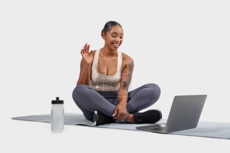 Photo for African American woman is seated on a yoga mat with a laptop in front of her. She is dressed in comfortable clothing suitable for yoga practice, have online fitness class - Royalty Free Image