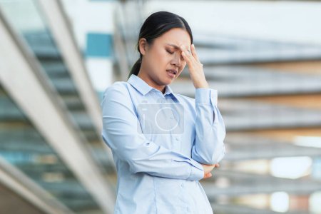 Asian woman in business attire appears distressed and holds her head, signaling a headache or overwhelming stress, against the backdrop of a modern urban environment
