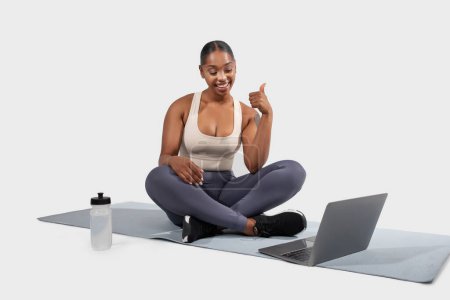 A radiant young African American woman sits cross-legged on a yoga mat, actively engaging with a virtual instructor on her laptop, white studio background