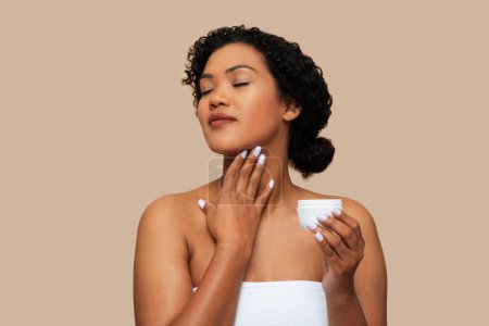A young woman with curly hair, wrapped in a white towel, gently applies moisturizing cream to her neck, moment of self-care and relaxation. The neutral beige background