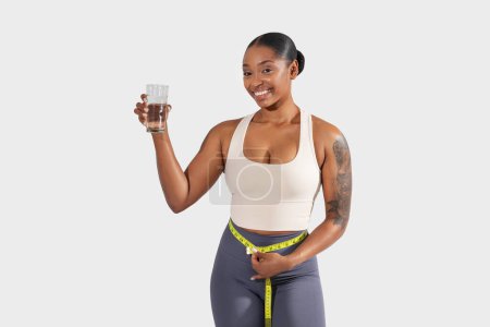 Une jeune femme noire heureuse se tient avec confiance avec un ruban à mesurer autour de sa taille, tenant un verre d'eau claire. Ses vêtements de sport suggèrent un accent sur la santé et la forme physique