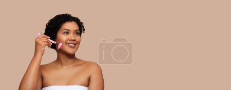 Beautiful young Brazilian lady wrapped in towel using pink face roller over beige studio background, looking at copy space, web-banner