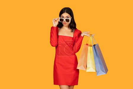 Eine Frau in einem leuchtend roten Kleid steht da und hält mehrere Einkaufstüten in den Händen. Taschen, die sie in der Hand hält, deuten darauf hin, dass sie Dinge eingekauft hat, ihre Sonnenbrille angepasst hat