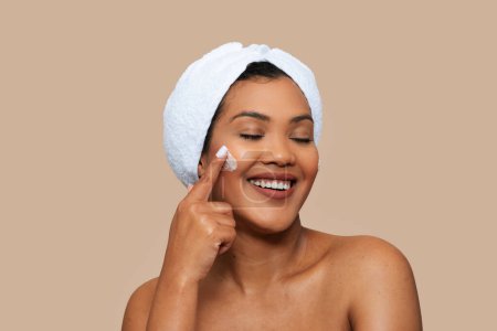 Une femme avec une serviette enroulée autour de sa tête applique doucement de la crème sur son visage. Elle se tient debout devant un miroir, se concentrant sur sa routine de soins de la peau.