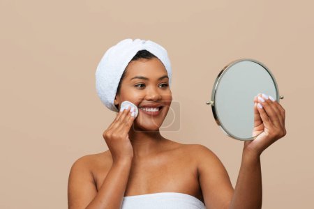 Eine fröhliche junge Frau, in ein weißes Handtuch gehüllt, betrachtet ihr Spiegelbild in einem Handspiegel, während sie Tonner auf ihre Wange aufträgt. Ihr Haar steckt unter einem weißen Stirnband
