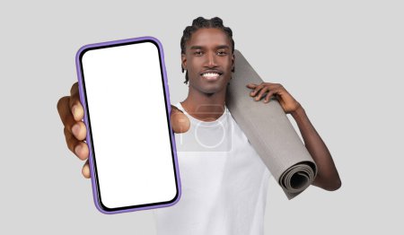 Black man standing while holding a yoga mat in one hand and a cell phone in the other. He appears to be preparing for a workout or fitness session.