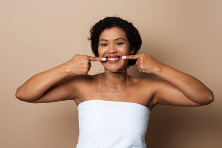 A cheerful young woman wrapped in a towel is demonstrating proper dental care by holding fingers to her mouth. She stands against a plain background, promoting healthy habits with a radiant smile.