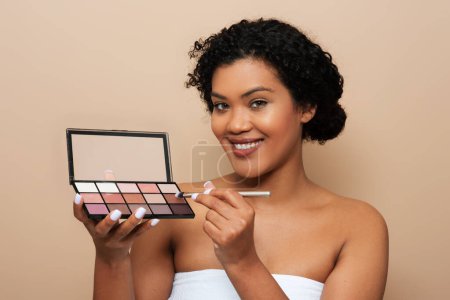 A young woman is shown holding a colorful makeup palette in her hand, examining the different shades and products. She appears focused on selecting the right colors for her look.