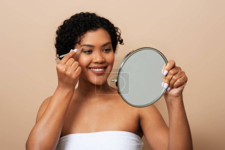 Eine junge hispanische Frau steht vor einem Spiegel und schminkt sich vorsichtig ihr Gesicht. Sie ist fokussiert und präzise und nutzt verschiedene Werkzeuge und Produkte, um ihre Merkmale zu verbessern.