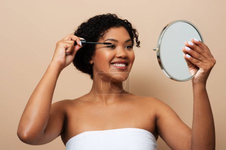 Foto de Una joven sonriente con el pelo rizado se está poniendo rímel en las pestañas mientras se mira en un espejo de mano. Ella está envuelta en una toalla blanca o tela - Imagen libre de derechos