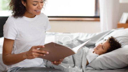 Una mujer afroamericana sonriente está sentada junto a una cama acogedora, absorta en la lectura de un cuento de un libro a su hijo pequeño, que está acostado, apareciendo contento y atento